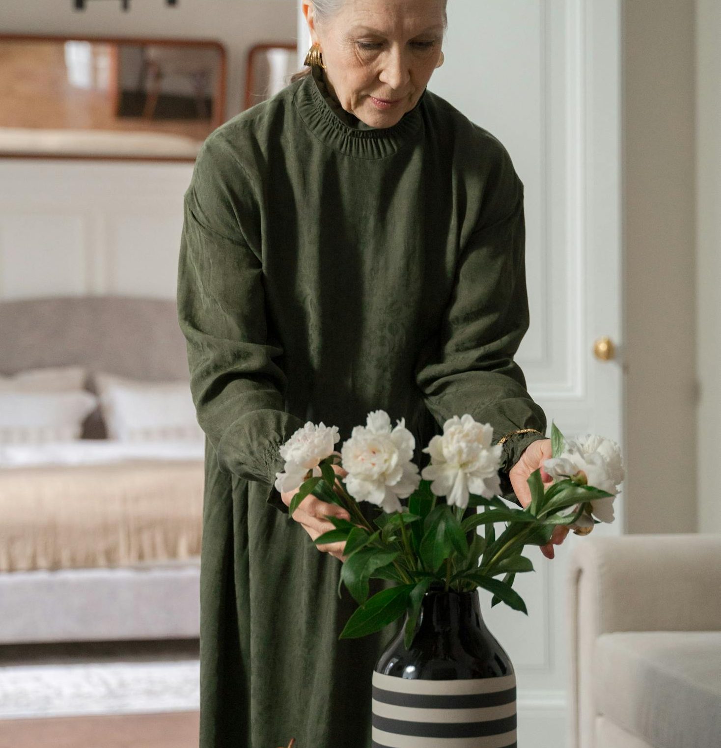 An elderly woman in a green dress arranges white peonies in a cozy living room, symbolizing tranquility.